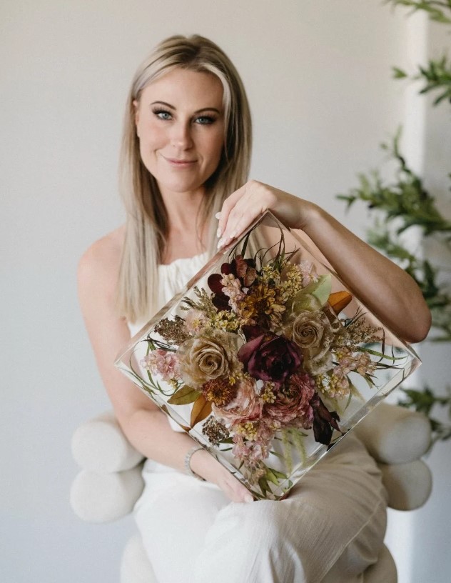 Crystal of Ever After bouquet holding a resin keepsake with preserved wedding flowers in shades of burgundy, cream, and pink, seated on a white chair in a bright room.