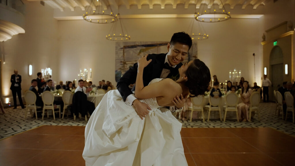 Nathan dipping Gabrielle during their first dance in the elegant ballroom, with chandeliers overhead and guests watching from round tables. Dreamwood Films with Stephy Plechas.