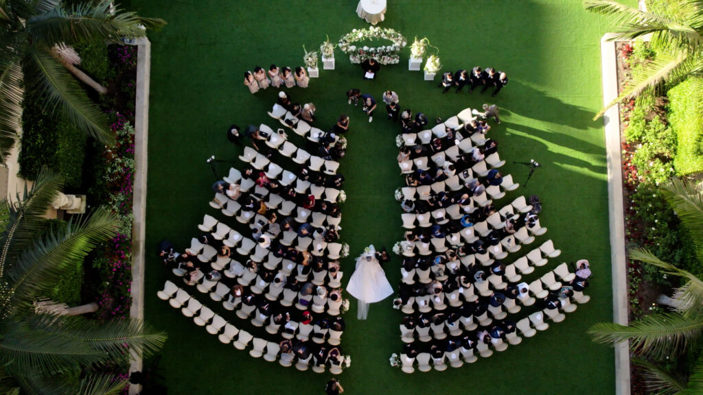 Aerial view of Gabrielle and Nathan’s wedding ceremony at The Maybourne Beverly Hills, with the bride walking down the aisle between curved rows of white chairs, a floral arch at the altar, and palm trees surrounding the lawn. Dreamwood wedding photography