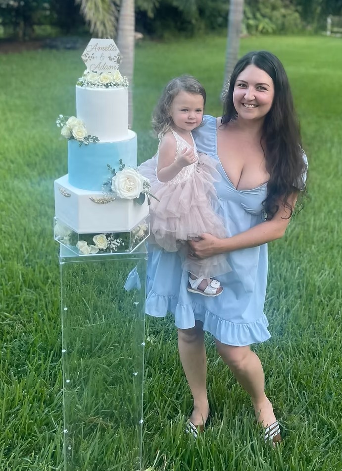 Heather Krevoy, owner of Brookie Cookie Bakery, holding a young child beside a custom three-tier white and light blue wedding cake with floral accents and a personalized cake topper on an acrylic stand outdoors.