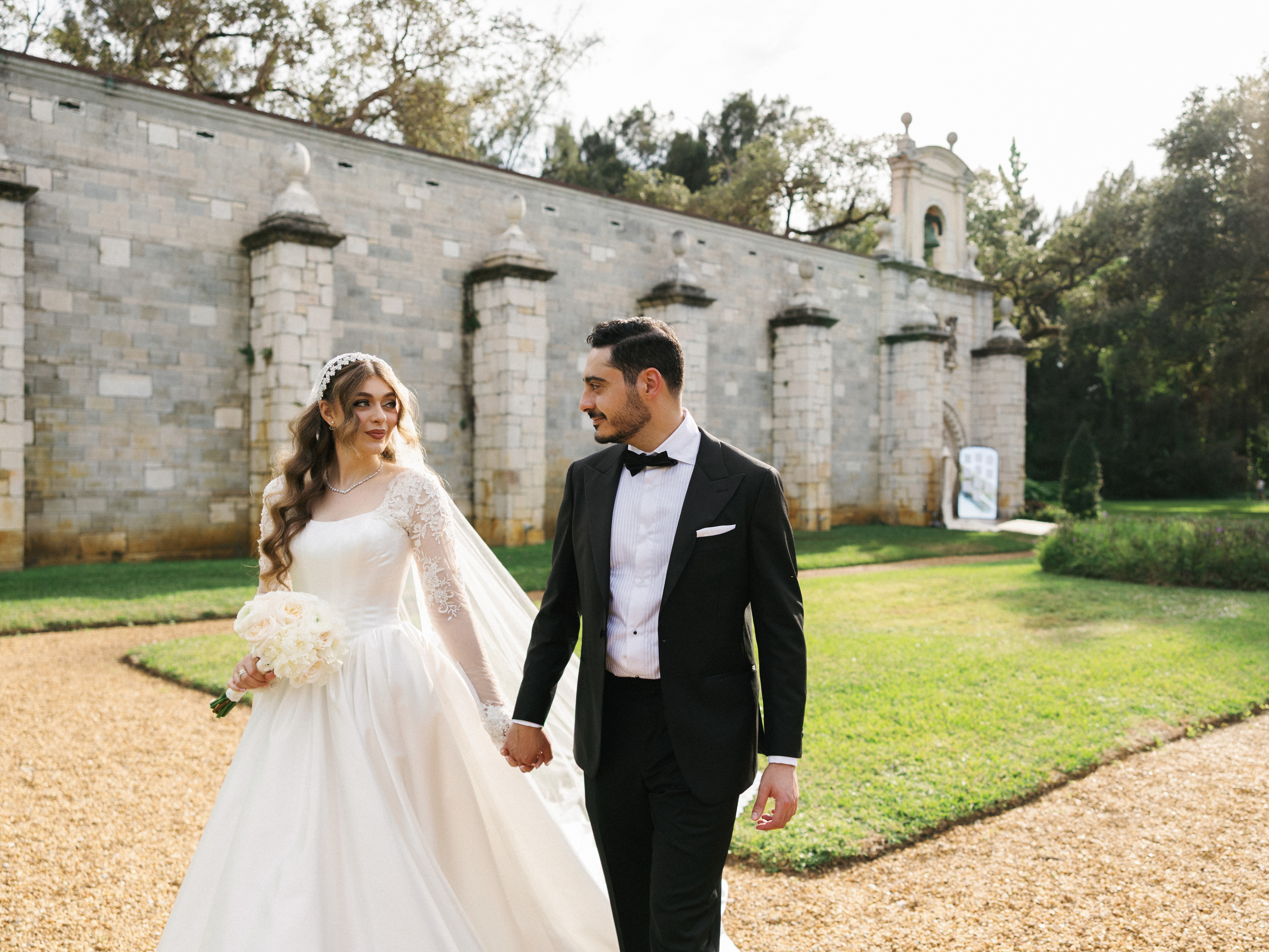 A bride and groom smiling at each other as they walk hand in hand along a gravel path in front of a historic stone building with a bell tower.