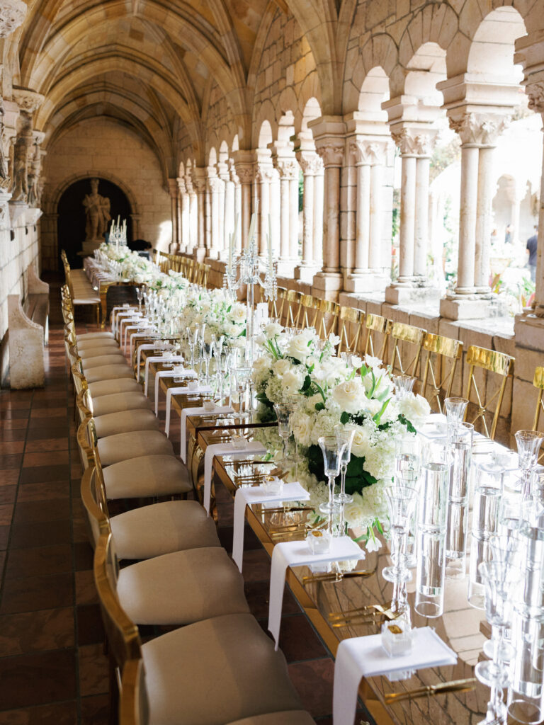 A long banquet table set for a wedding reception inside a historic stone corridor with arched columns, decorated with white floral centerpieces, gold chairs, and tall glassware.