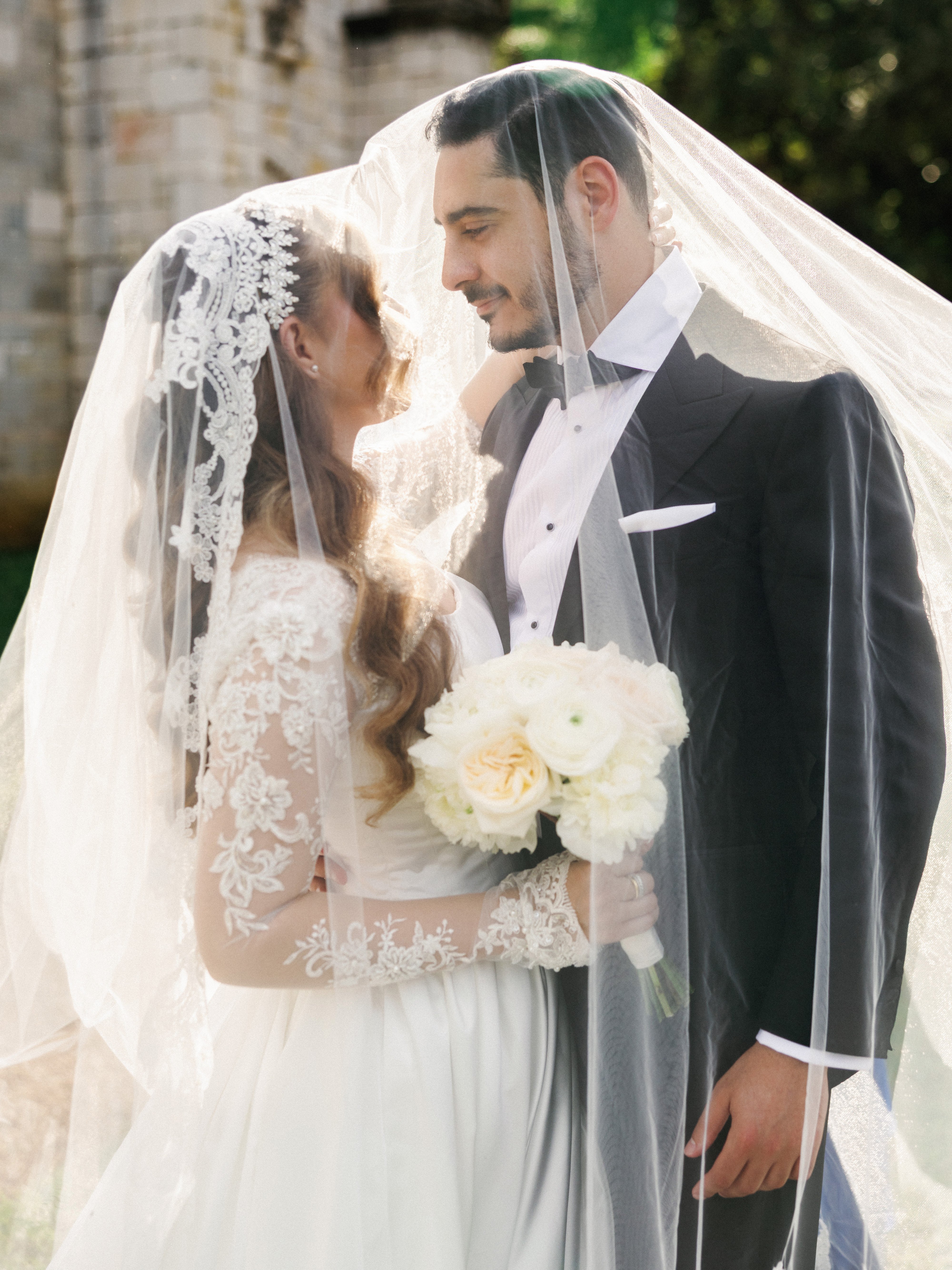 A close-up portrait of a bride and groom looking into each other's eyes under a sheer, sunlit veil, with the bride holding a white floral bouquet.
