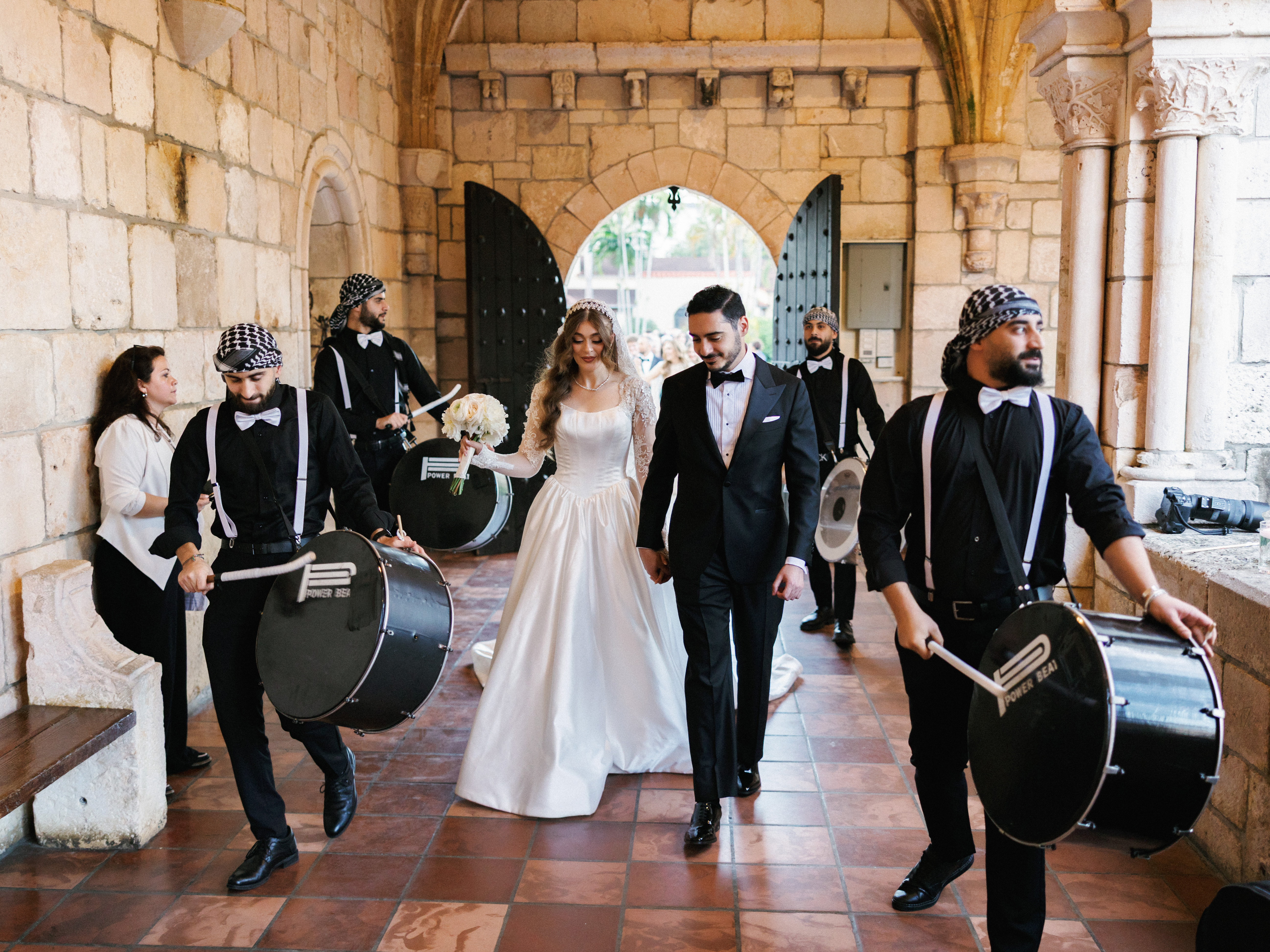 A bride and groom walking through a historic stone archway, surrounded by drummers in black outfits and white suspenders playing celebratory drums.