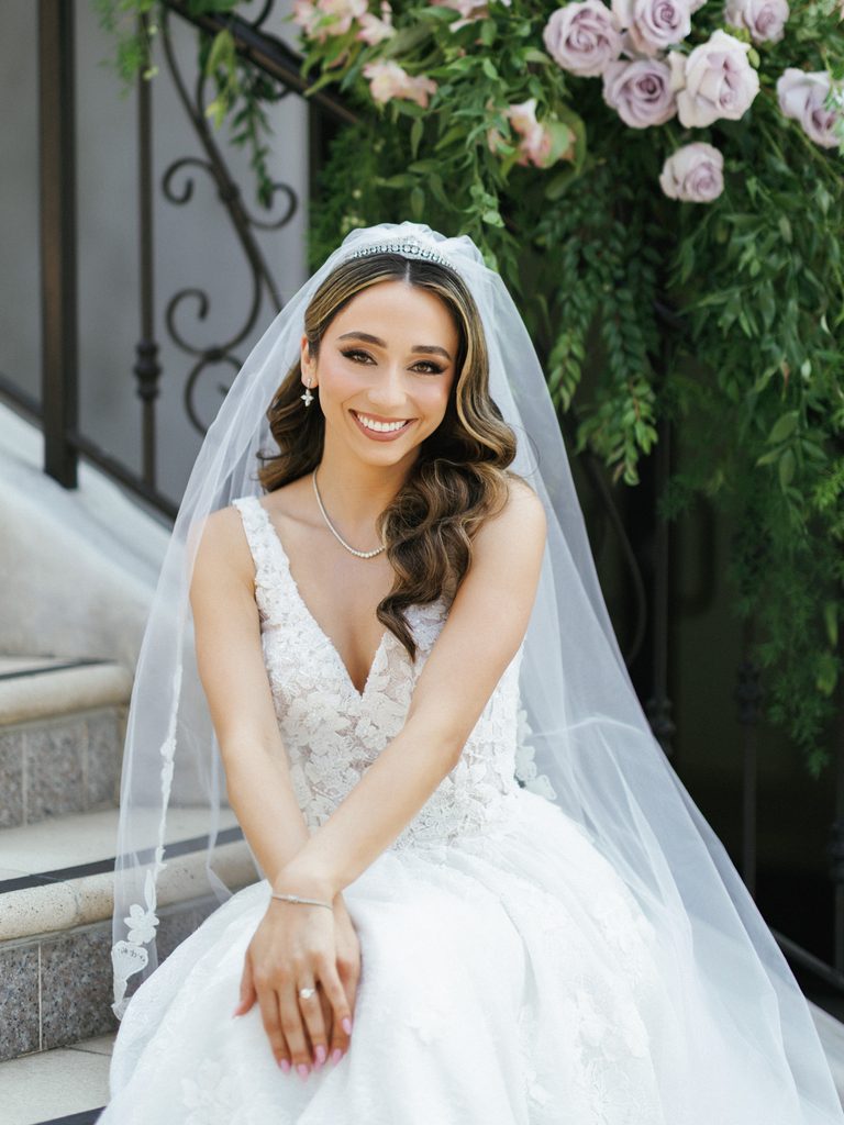 Happy bride with elegant wavy hair and long tulle veil looking directly at camera outdoors