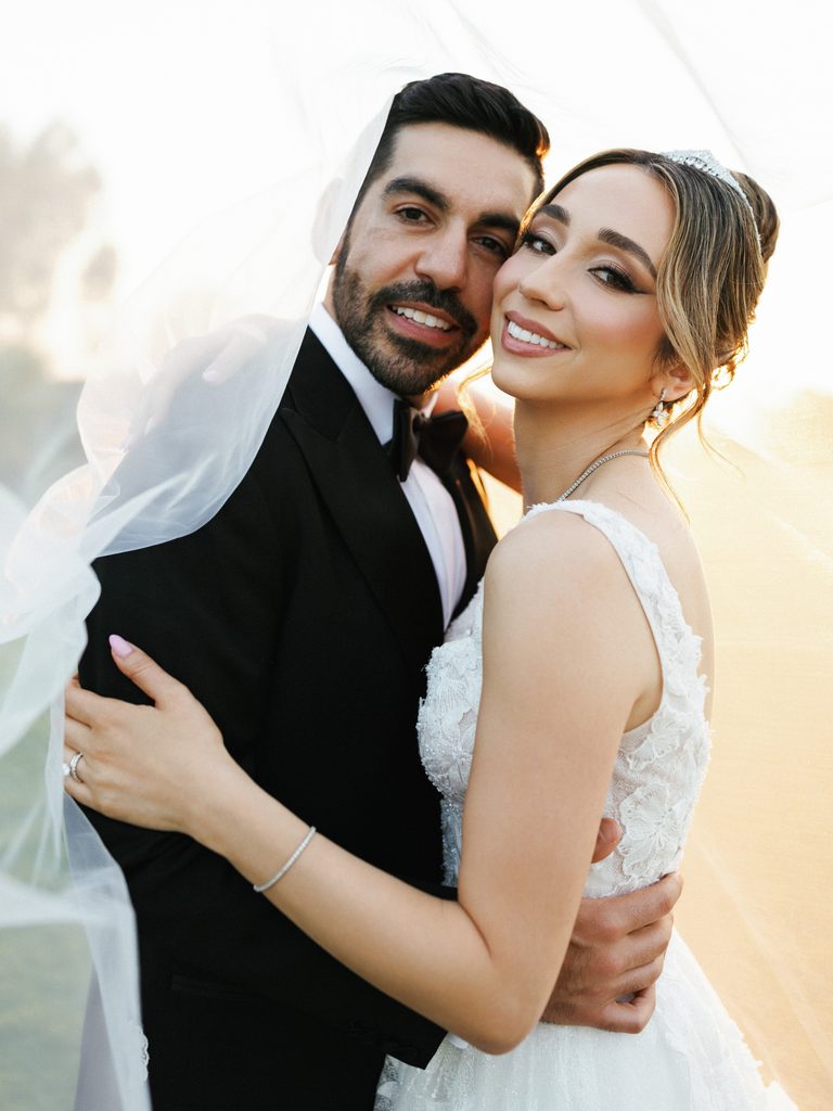 Beautiful smiling bride and groom looking at camera wrapped in long white tulle veil