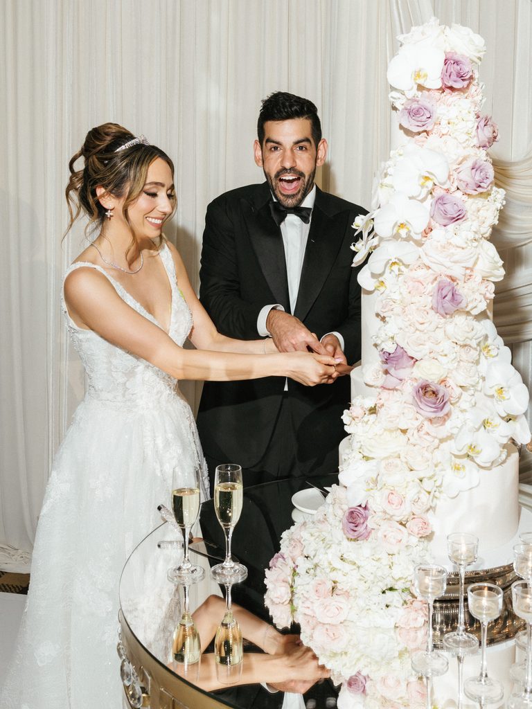 Excited groom making a funny face while bride cuts huge luxury wedding cake decorated with cascading roses