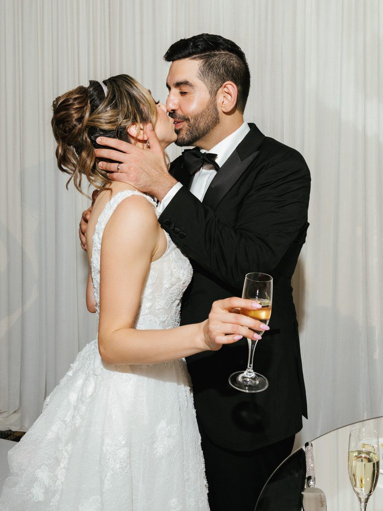 Elegant newlyweds embracing and sharing a sweet kiss while celebrating with sparkling wine at banquet