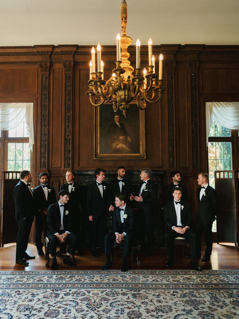 Wide angle shot of groomsmen gathering in luxury estate ballroom before wedding ceremony