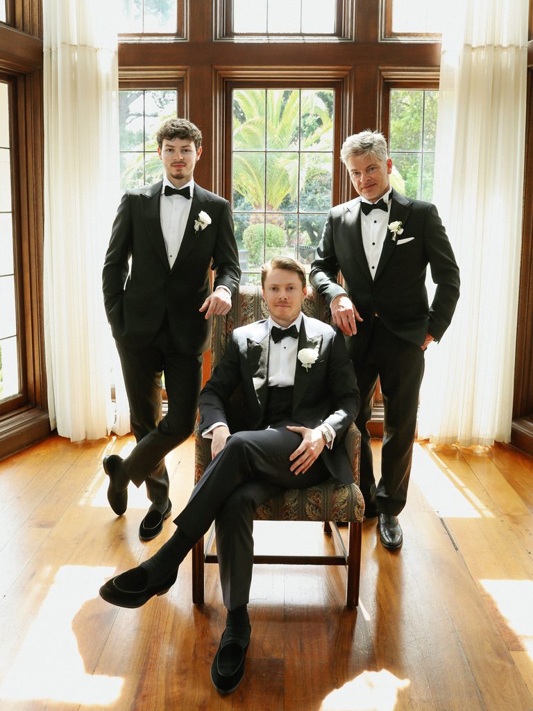 Handsome groom sitting in vintage chair with groomsmen in classic black tuxedos near large window