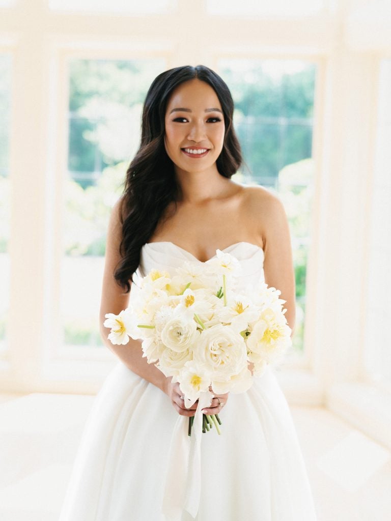 Smiling bride in strapless dress holding white peony and garden rose bouquet indoors