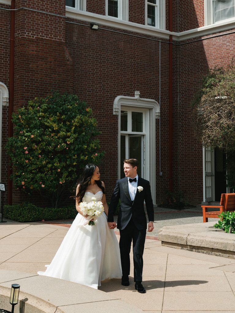 Elegant newlyweds holding hands while walking near historic brick mansion architecture