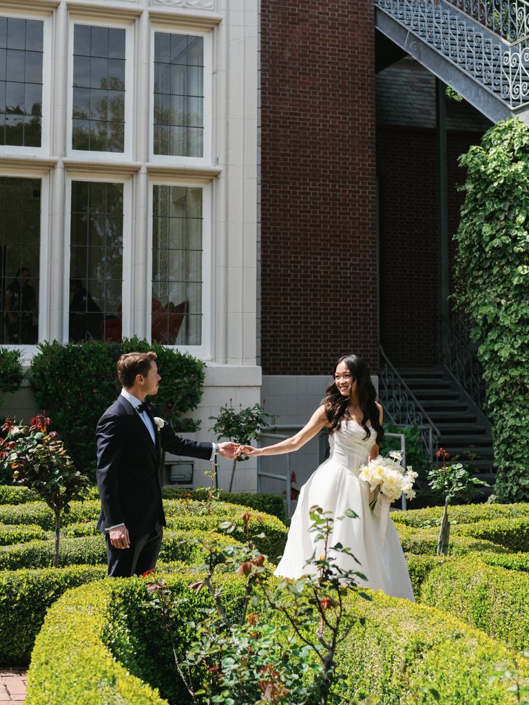 Elegant newlyweds strolling between manicured green hedges outside historic brick estate windows
