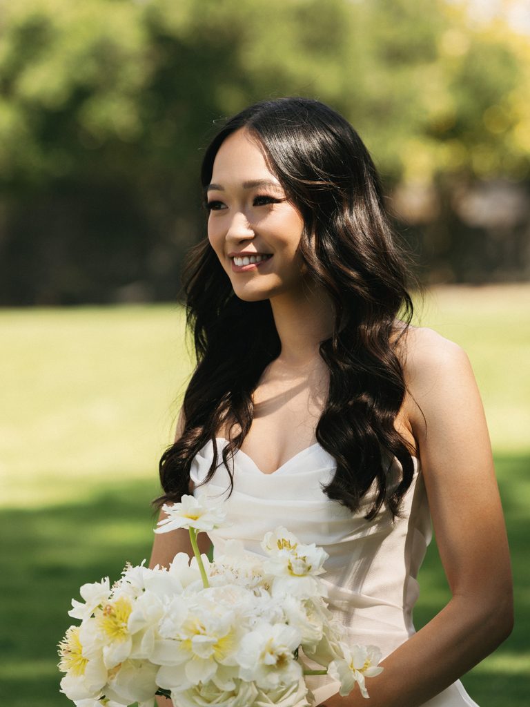 Beautiful bride with long wavy hair smiling outdoors while holding white wedding bouquet