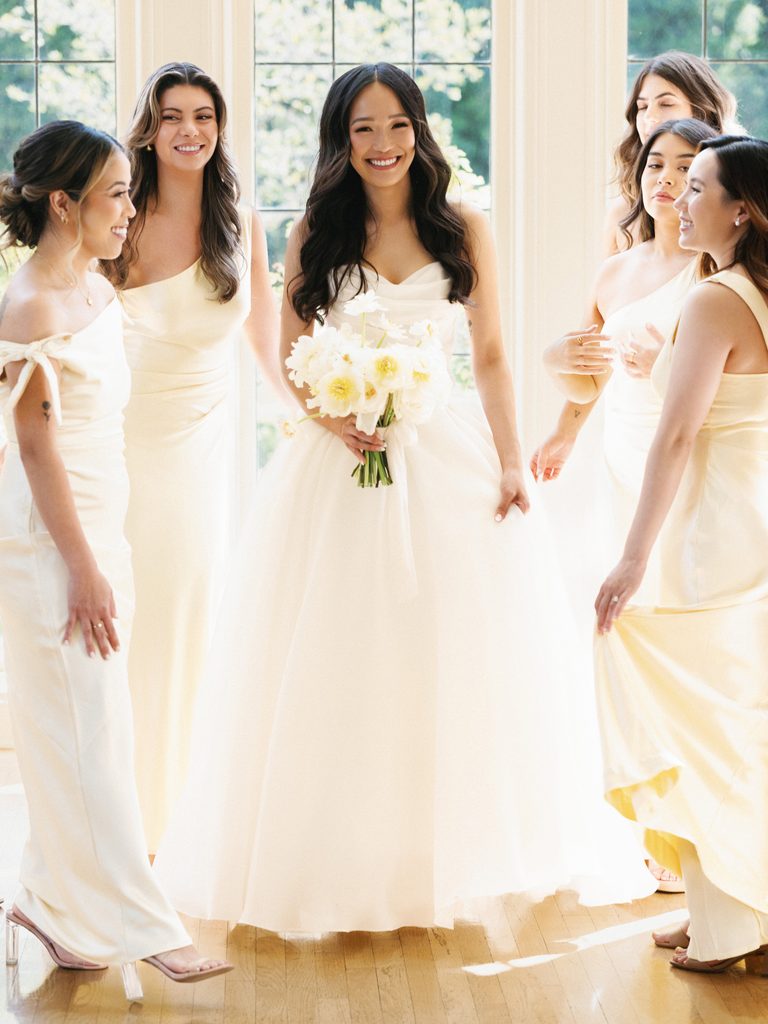 Elegant bridesmaids wearing chic pale yellow gowns standing around smiling bride with white flowers