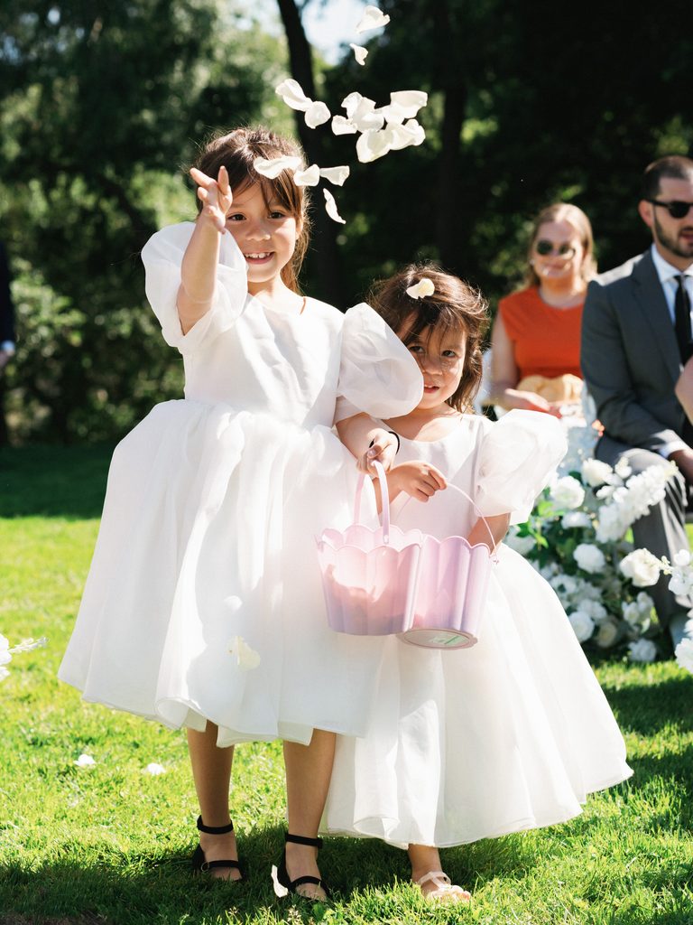 Adorable flower girls in fluffy white dresses throwing petals down the aisle during outdoor wedding ceremony