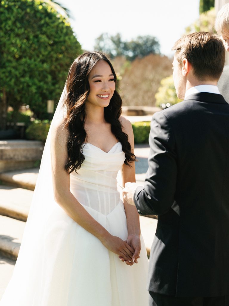 Smiling bride wearing long tulle veil and strapless wedding dress looking at groom during outdoor ceremony