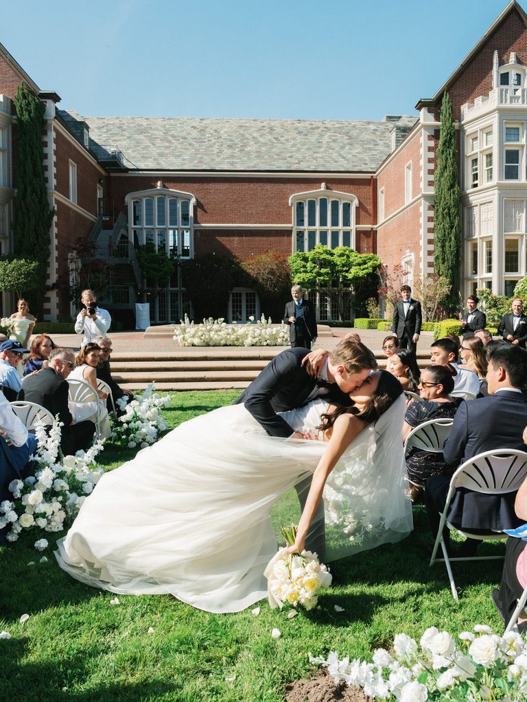 Passionate bride and groom kissing on green lawn with applauding wedding guests in background