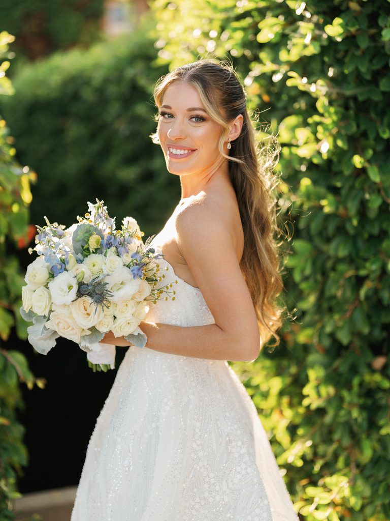 Smiling bride with long wavy hair holding white bouquet outdoor at luxury vineyard wedding