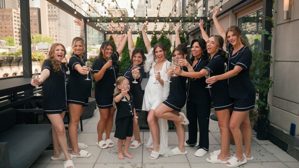 Wedding morning. Sammi celebrating her getting-ready moments on a rooftop terrace with her bridal party. They are raising champagne glasses and cheering with a New York skyline in the background. This fun wedding celebration was documented by Dreamwood Films.