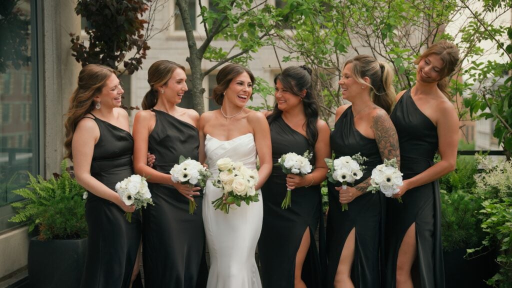 Sammi sharing a joyful laugh with her bridesmaids, who are wearing elegant black one-shoulder dresses and holding white bouquets. The bride's hair is beautifully styled by Yuliia Zablotska, captured outdoors by Dreamwood Films during a wedding celebration.