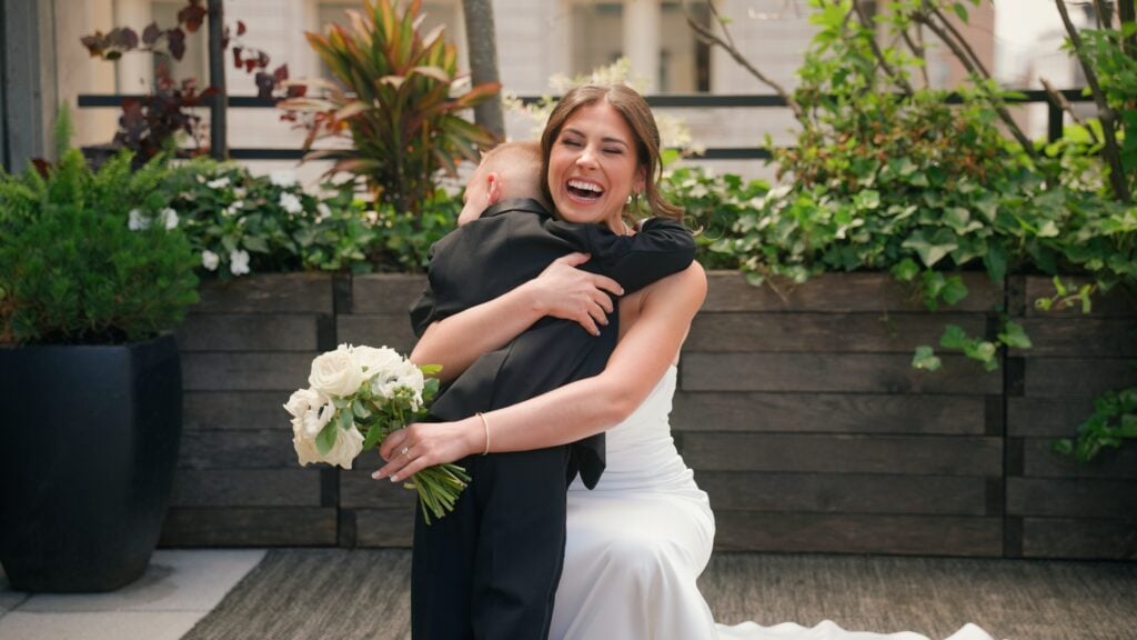 A joyful bride kneeling on a balcony to hug a young boy in a black suit, laughing brightly while holding her white rose bouquet. Her hair is beautifully styled by Yuliia Zablotska, capturing the pure happiness of this wedding celebration. Dreamwood Films.