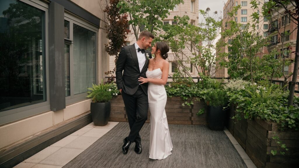 A bride and groom smiling at each other on a New York City rooftop terrace surrounded by lush greenery and city buildings. The bride's elegant hair and dress look stunning in this candid portrait captured by Dreamwood Films.