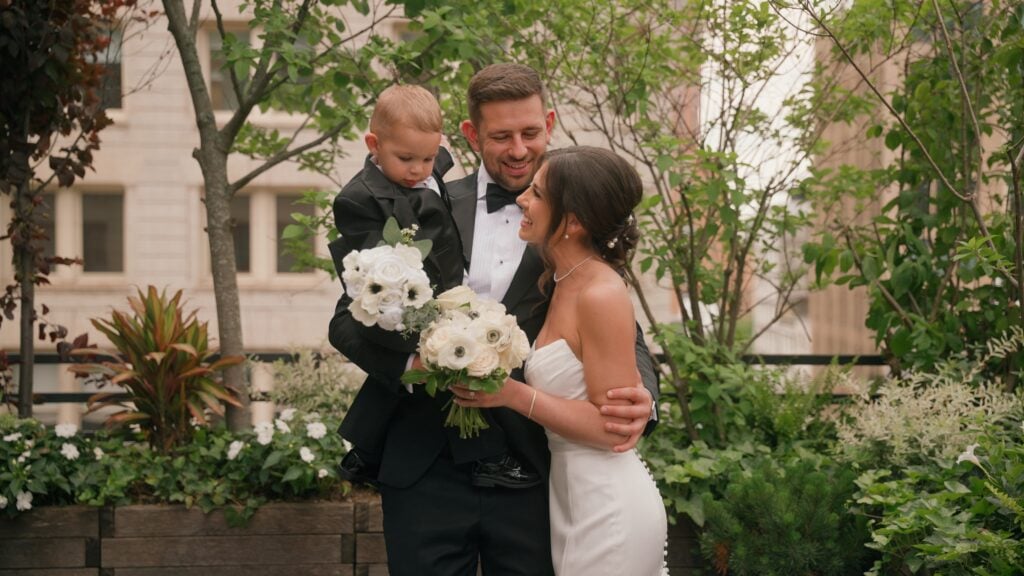 A groom in a black tuxedo holding a young boy in a matching suit while the bride, wearing a strapless white gown and a timeless updo styled by Yuliia Zablotska, smiles warmly at them. This sweet family moment was captured on a New York City rooftop by Dreamwood Films.