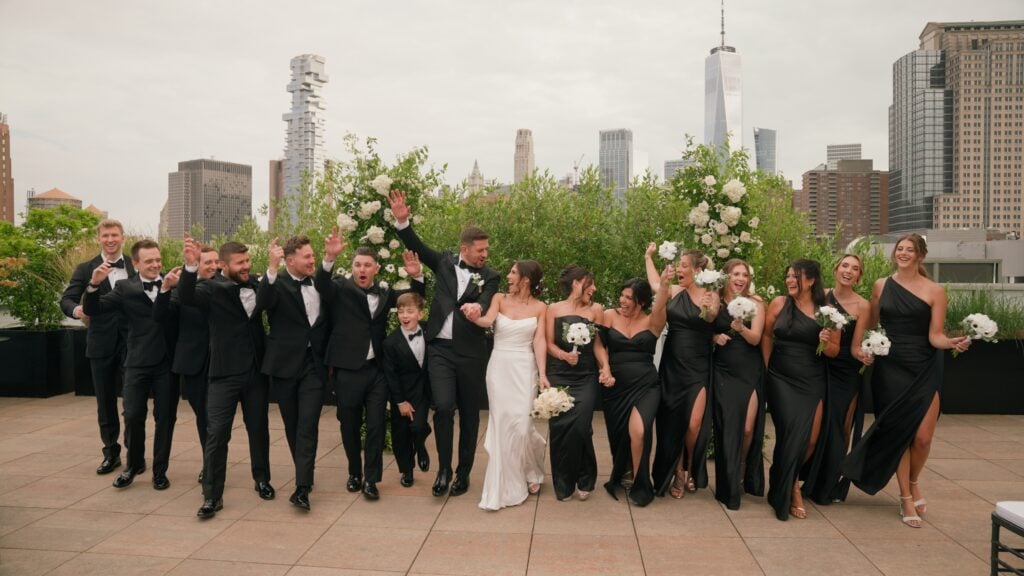 A large wedding celebration on a rooftop in New York. The bride and groom walk hand-in-hand, surrounded by cheering bridesmaids in black gowns and groomsmen in black suits, with their arms raised in the air. A beautiful moment documented by Dreamwood Photo, featuring hair styling by Yuliia Zablotska.
