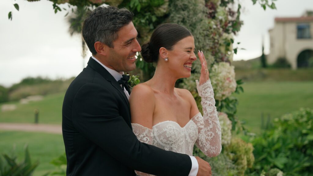 A groom hugging his bride as she laughs joyfully, with soft green and white wedding florals in the background. Dreamwood films.