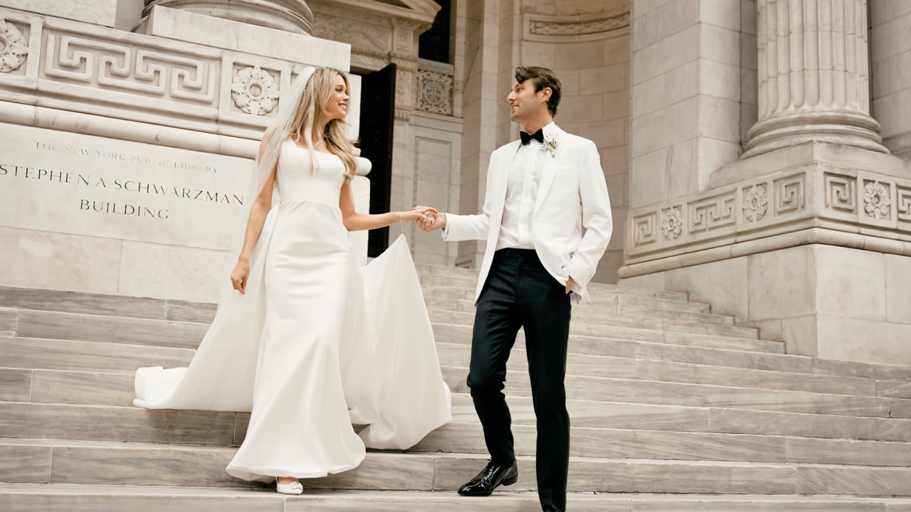 Frame from a wedding film: bride and groom holding hands on broad stone steps beside carved columns.