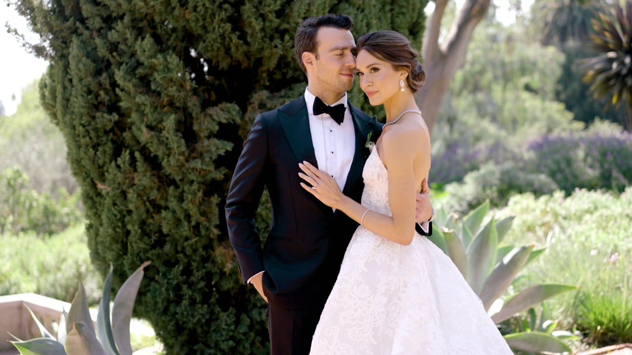Frame from a wedding film: bride and groom standing close together among trees and garden plants.