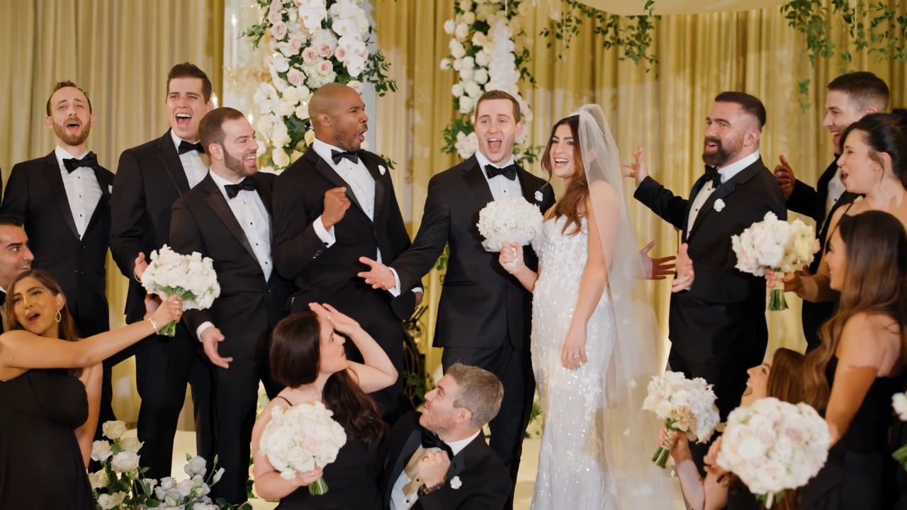 Frame from a wedding film: bride, groom and wedding party with white bouquets in front of gold curtains.