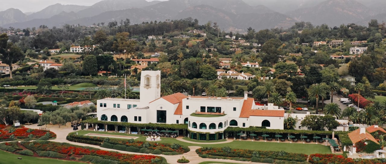 Frame from a wedding film: aerial view of a white building with red-tiled roofs, formal gardens and mountains beyond.