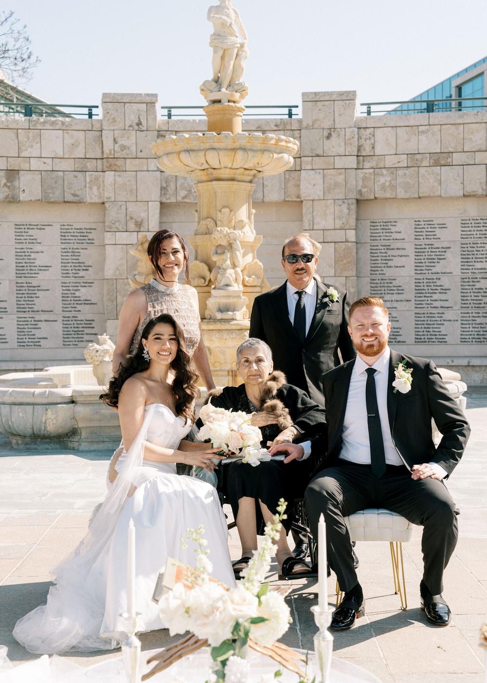 Bride and groom seated beside an older woman, with two people standing behind them by a stone fountain.