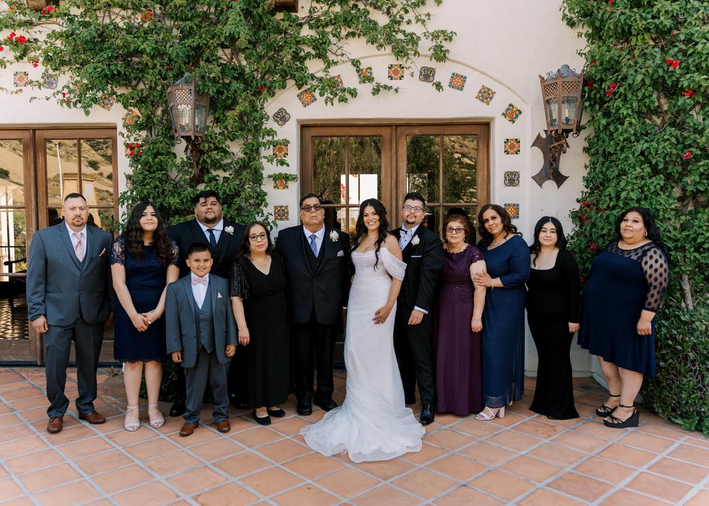 Bride, groom and a wedding party posing under flowering vines beside a white building.