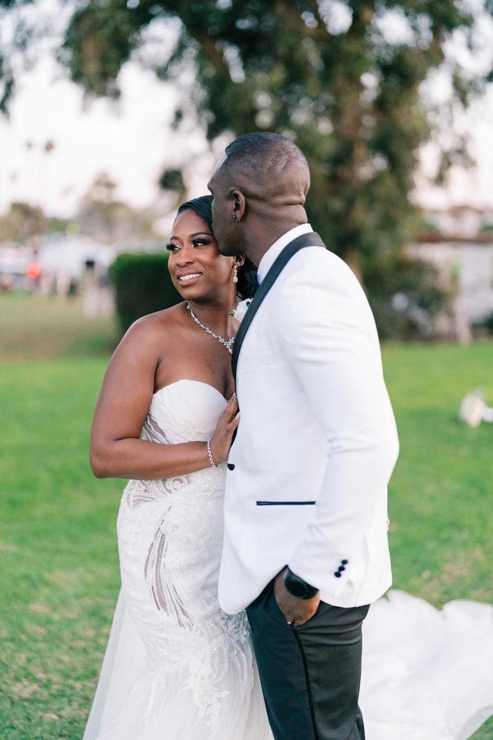 Bride smiling beside a groom in a white jacket on a lawn.